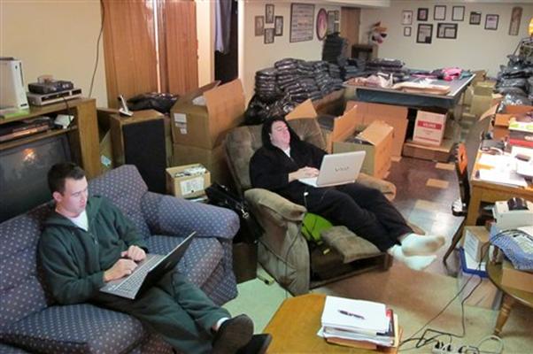 Two young men dressed in sweatpants, sitting on a couch and recliner, working on laptops in a cluttered room.