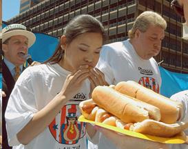 Young woman eating a hot dog as part of hot dog eating contest with a plate full of hot dogs in front of her.